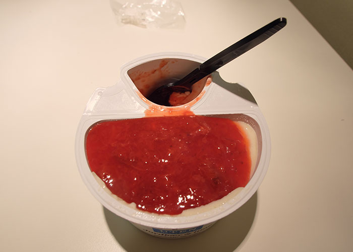 Plastic container of strawberry yogurt with a black plastic spoon, illustrating food thieves at work in the office.