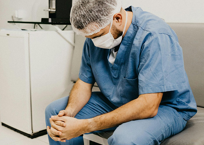 Healthcare worker in scrubs and mask sitting with head down, reflecting on things people believed were normal until truth revealed