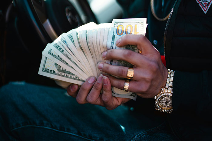 Person wearing gold jewelry holding a large stack of cash, illustrating ultra rich individuals and their lifestyle.
