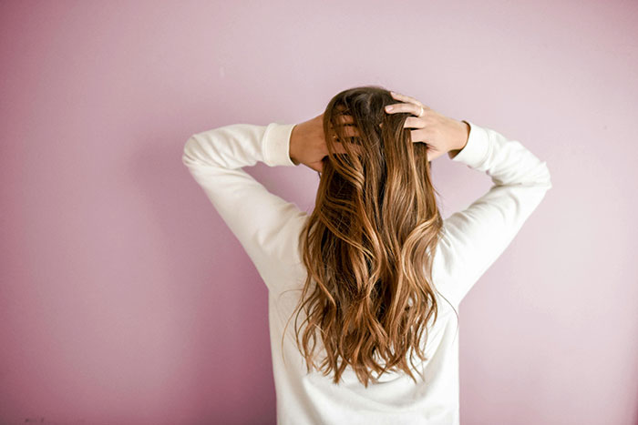 Woman with long wavy hair in white sweater, posing against a pink background representing ultra rich individuals lifestyle.