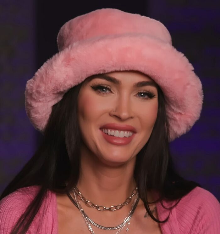 Smiling woman in a pink fluffy hat and matching sweater against a dark background.