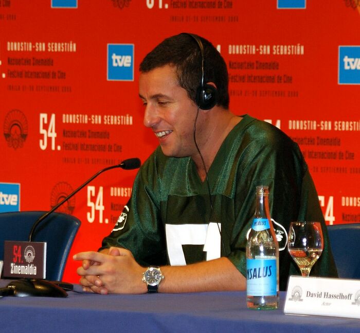 Actor speaking at a film festival, wearing a headset and green jersey, with a red backdrop and microphones.