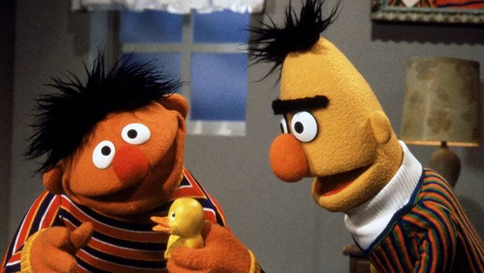 Iconic duo with colorful puppets, one holding a rubber duck, in a cozy room.