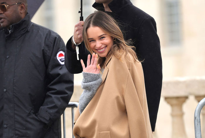  A B list celebrity, Emilia Clarke, is smiling and waving while wearing a beige coat and gray sweater, standing outdoors with security personnel nearby.
