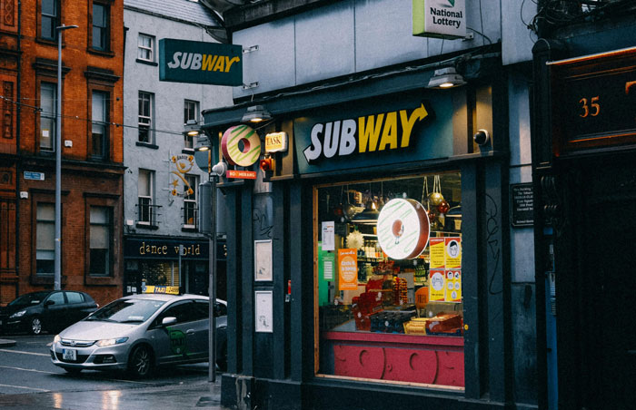 Subway restaurant on a rainy urban street corner with visible signage and nearby parked car at dusk