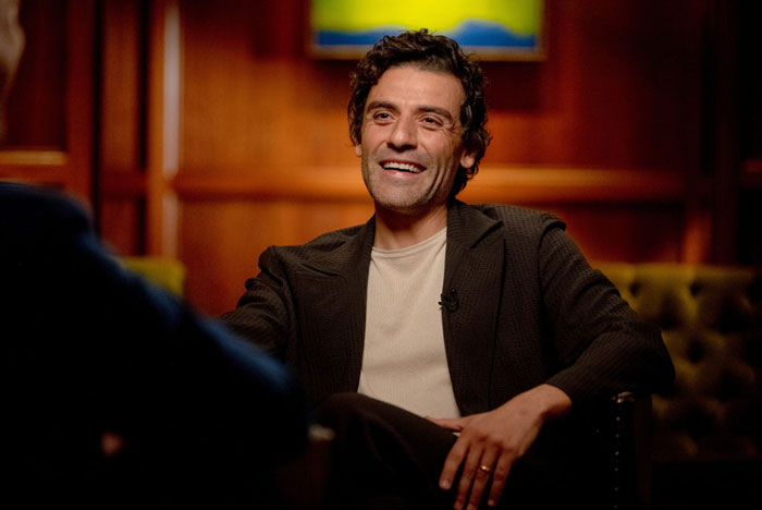  A B list celebrity, Oscar Isaac, is seated in an interview setting, smiling while wearing a dark blazer over a light-colored shirt in a warmly lit room.
