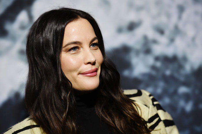  A B list celebrity, Liv Tyler, is smiling softly while wearing a striped sweater over a black top, with her dark hair flowing down at an indoor event.