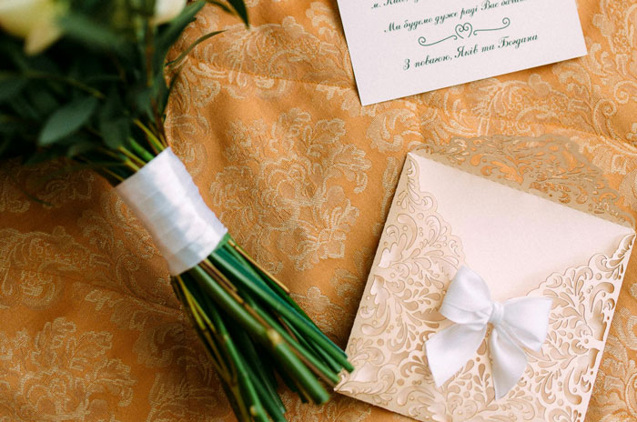 Bouquet wrapped in white ribbon next to an ornate invitation card with a bow on a patterned fabric background.