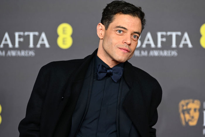  A B list celebrity, Rami Malek, is posing on the red carpet at the BAFTA Film Awards, wearing a dark suit, bow tie, and coat, with a branded backdrop behind him.