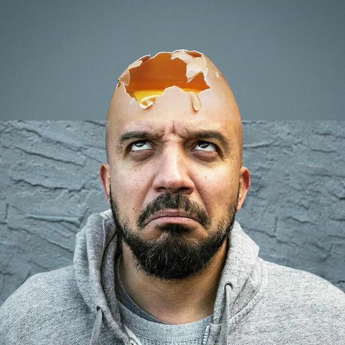 Man with an eggshell c*****d on his head, illustrating surrealism art, wearing a gray hoodie against a textured wall.
