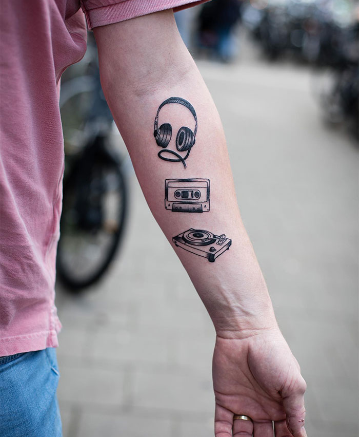 A minimalist tattoo on the forearm featuring a set of headphones, a cassette tape, and a turntable, representing a love for vintage music. Ideal tattoo ideas for men.