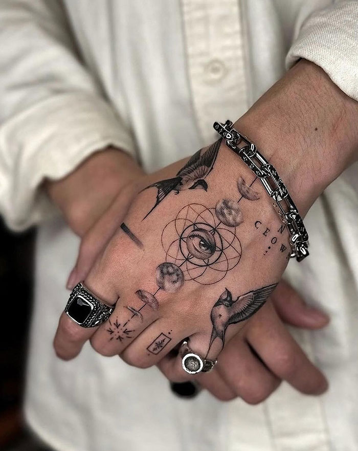  A detailed hand tattoo featuring symbolic elements like a central eye, geometric patterns, a bird, and celestial shapes, offering intricate and modern tattoo ideas for men.