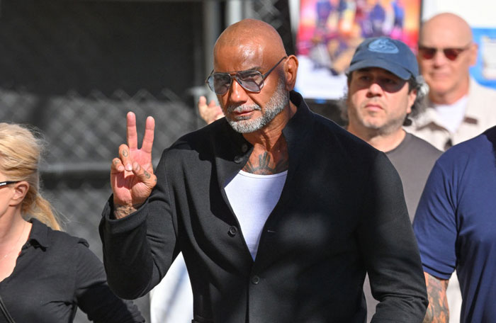  A B list celebrity, Dave Bautista, is walking outdoors, wearing a black jacket and glasses, flashing a peace sign with his fingers while surrounded by people.