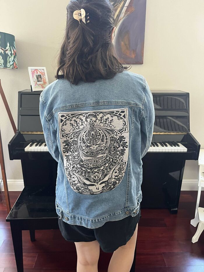 Person wearing a denim jacket with a visibly mended artistic patch standing in front of a black piano indoors.