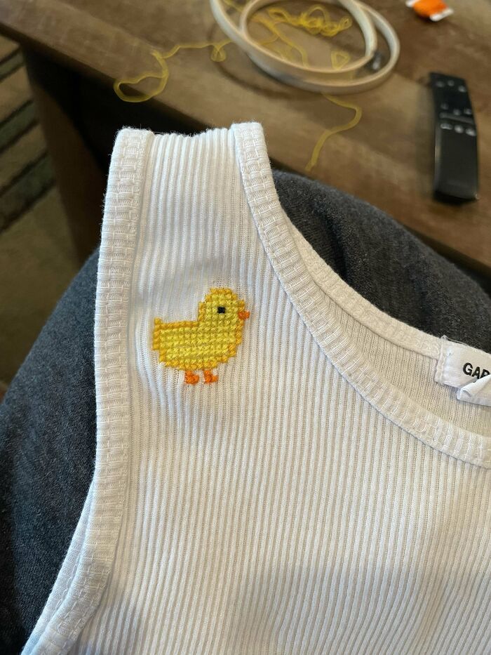 White ribbed tank top with a visible yellow cross-stitched duck patch mending a worn spot, showcasing creative visible mending.