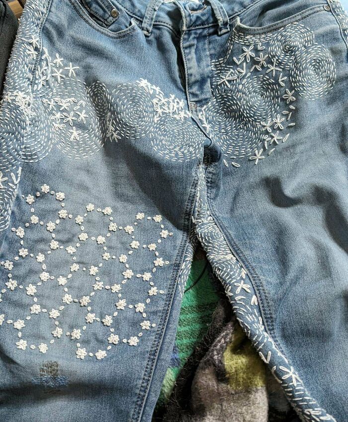 Blue jeans visibly mended with intricate white embroidery, showcasing creative and cool repairs on damaged clothing.