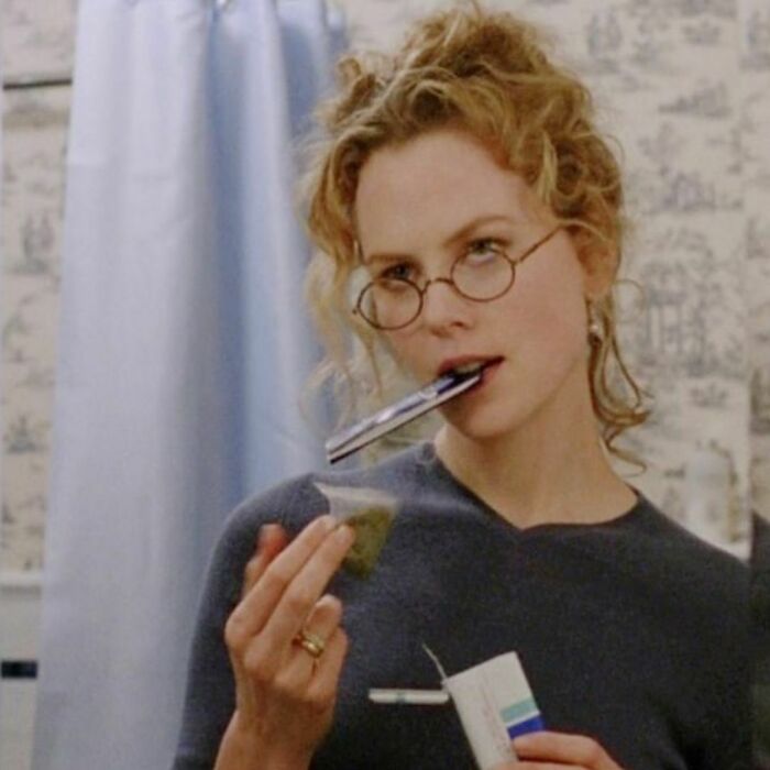 Woman with round glasses and curly hair holding a tea bag, embodying a '90s style moment.