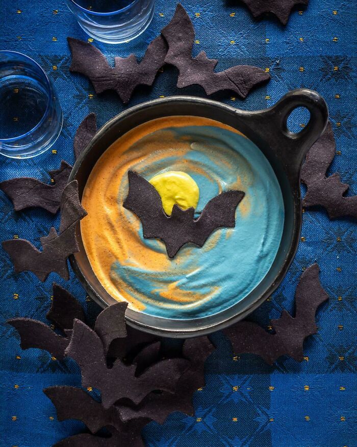 Halloween snacks featuring bat-shaped chips with colorful blue and orange dip in a black serving dish on a blue cloth.