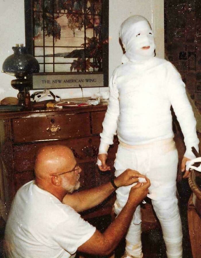 Child in mummy costume being helped by an older man, capturing a humorous blunder moment at home.