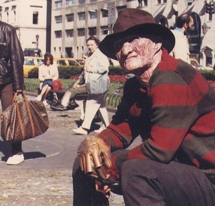 Man dressed as a horror character, wearing a striped sweater and hat, sits in a busy '90s city square.