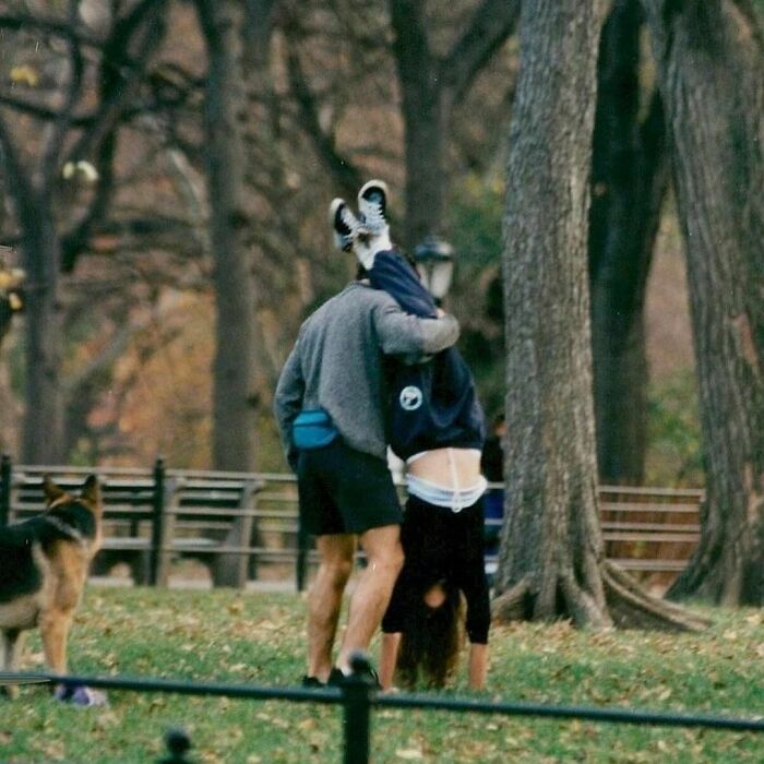 ‘90s moment: person playfully carrying another upside down in a park, with a dog watching nearby.