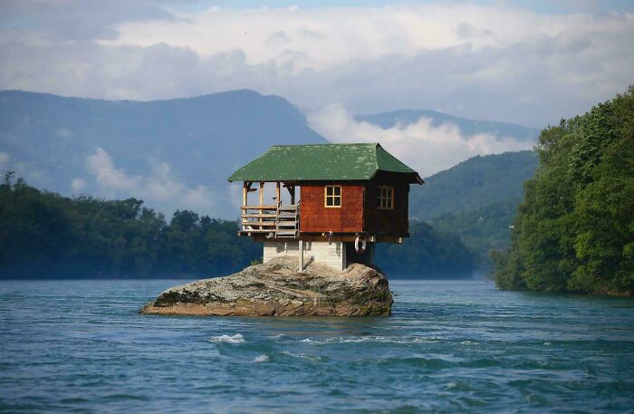 Unique bizarre building perched on a rock in the middle of a river with mountains and trees in the background