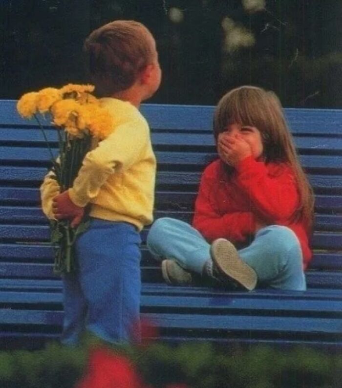 Young boy with hidden flowers on a bench, surprising a laughing girl in a nostalgic ‘90s moment.