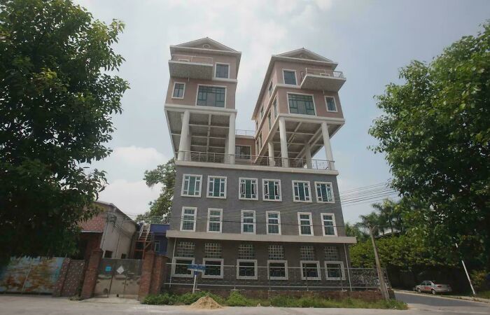 Unique bizarre building with upside-down houses elevated on stilts above a traditional multi-story structure surrounded by trees.