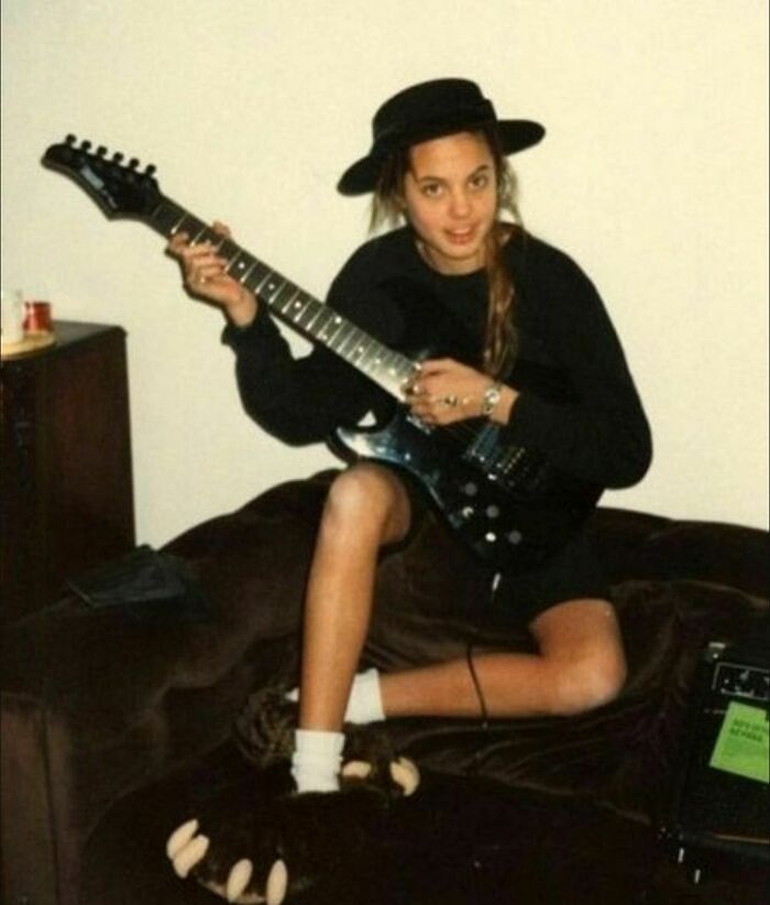 Person in the '90s holding an electric guitar, wearing a black hat and bear paw slippers, seated on a couch.
