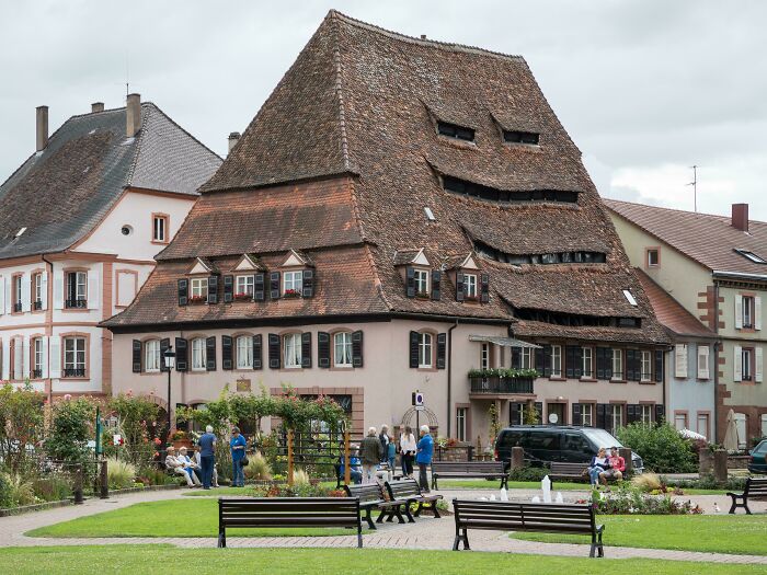 Bizarre building with a warped roof and windows resembling a smiling face, surrounded by people and benches in a park.