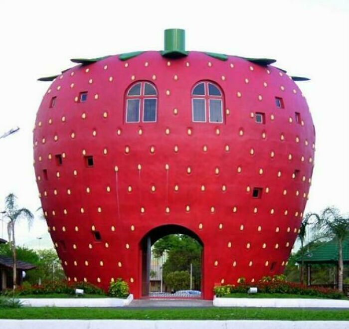 Bizarre building shaped like a giant strawberry with windows and an arched entrance surrounded by greenery.