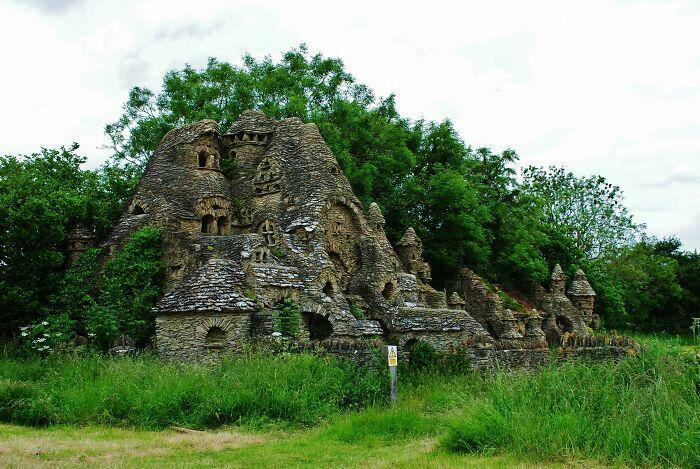 Stone bizarre building with unique castle-like features surrounded by lush greenery and tall grass under a cloudy sky