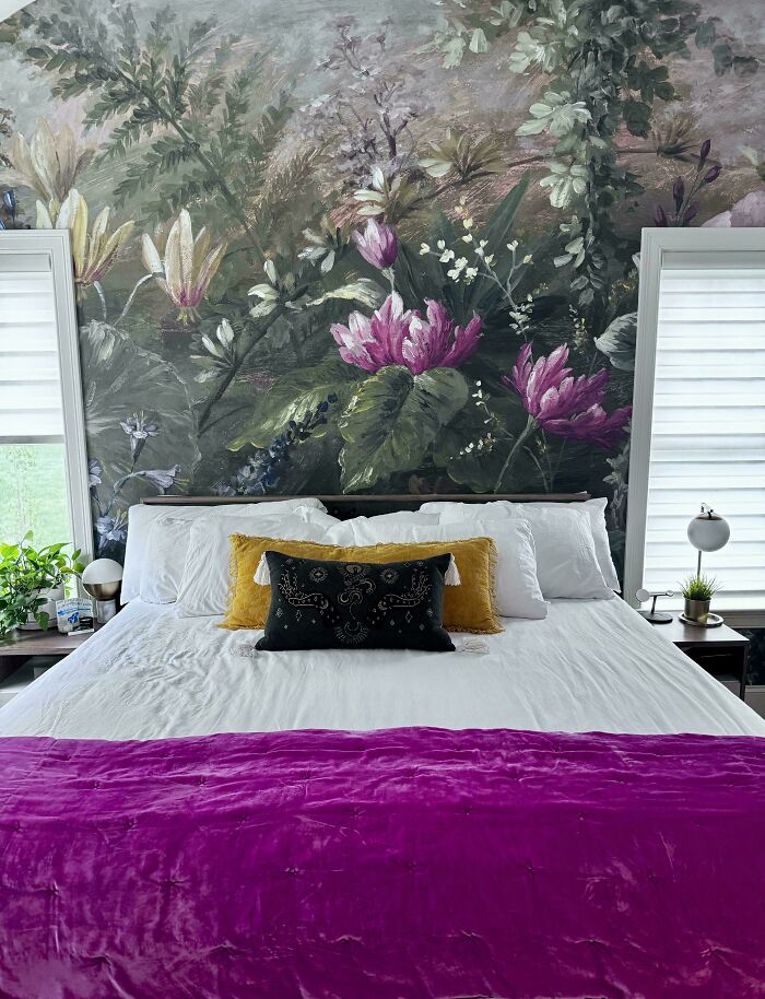 Bed seamlessly merges with floral mural background, with white bedding, gold and black pillows, and purple throw.