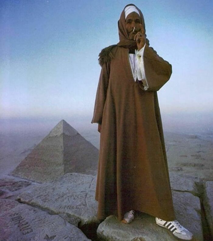 Man standing on a pyramid wearing traditional attire and Adidas sneakers, smoking with a scenic background. Pics That Go Hard.