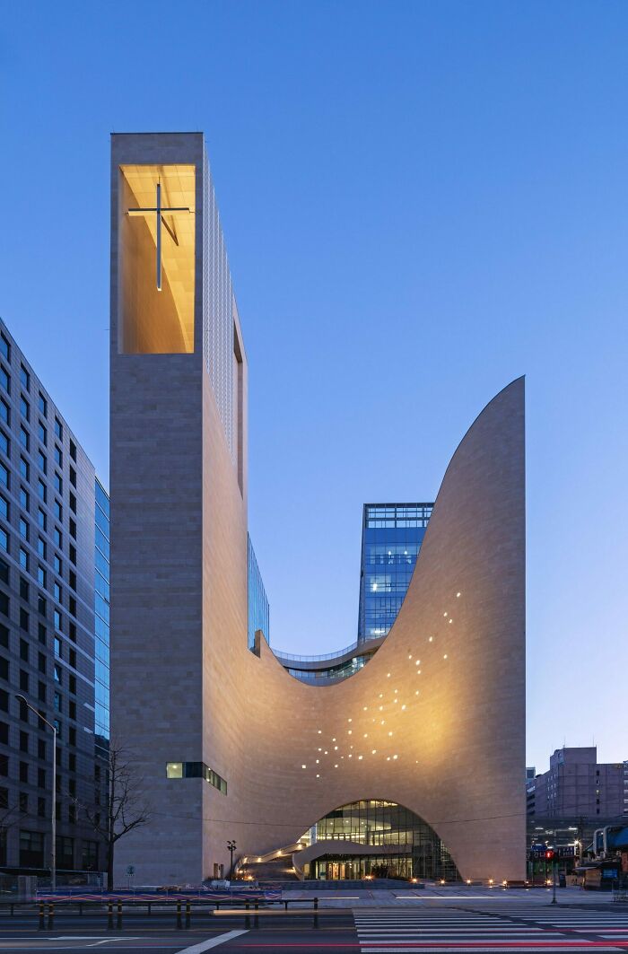 Modern bizarre building with unique curved design and glowing cross at dusk in urban setting with clear sky.
