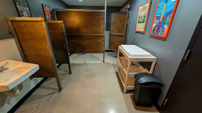 Baño público con divisores de madera y área de cambio para bebés en espacio limpio y moderno.