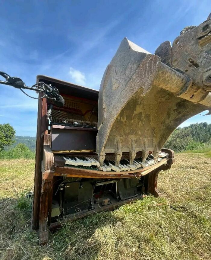 Excavator bucket smashing into a piano in a field under a clear blue sky, creating a dramatic visual impact, hard pic.