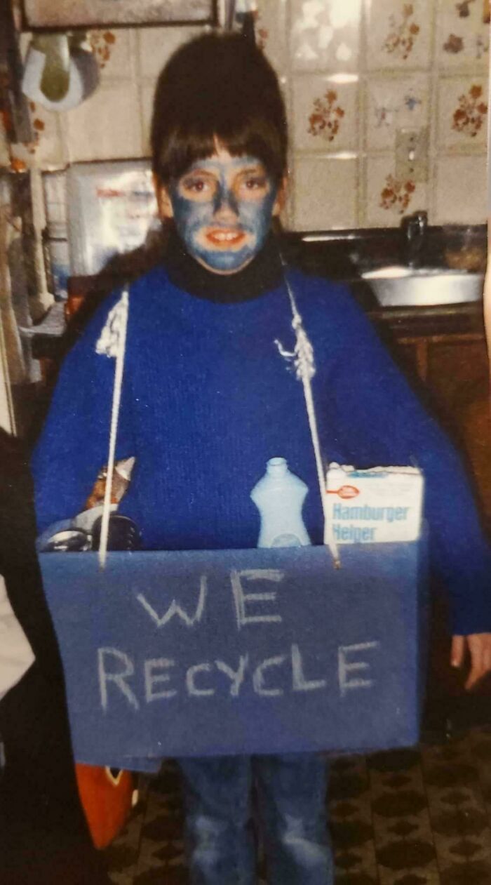 Child in blue face paint and costume with "We Recycle" sign, embodying retro blunder years fashion.