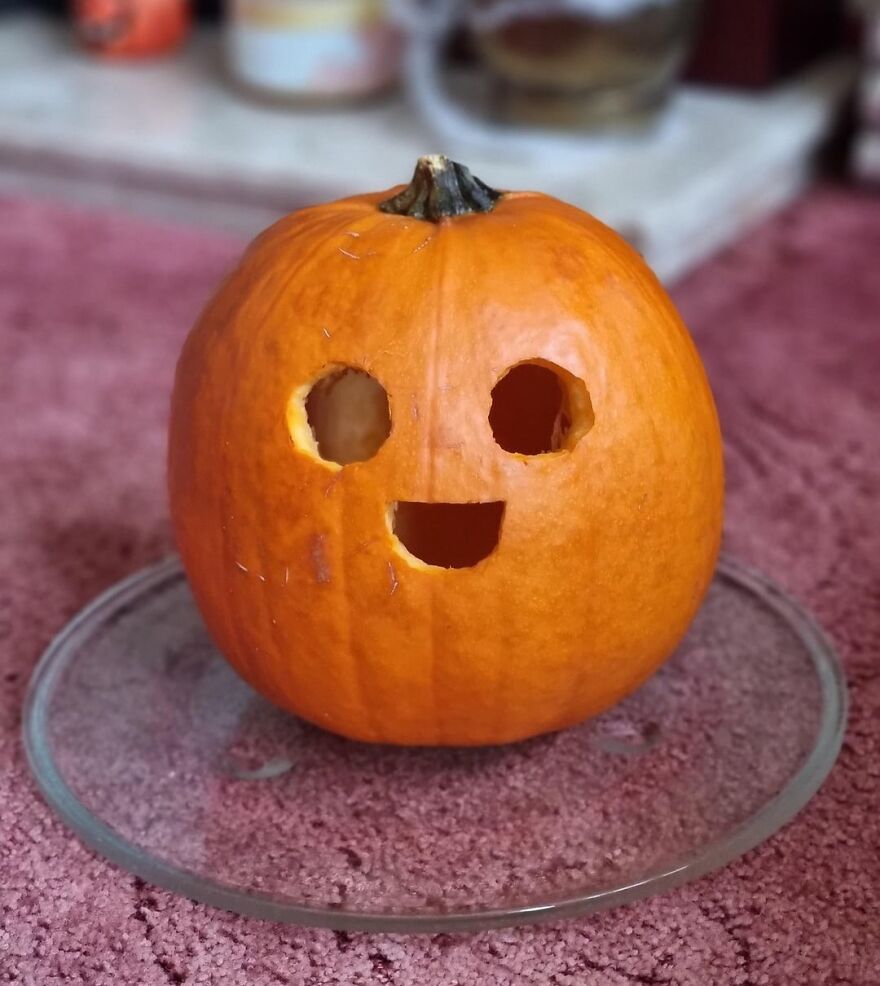 Small pumpkin carving with quirky smiling face, featuring simple round eyes and a wide mouth, displayed on a glass plate.