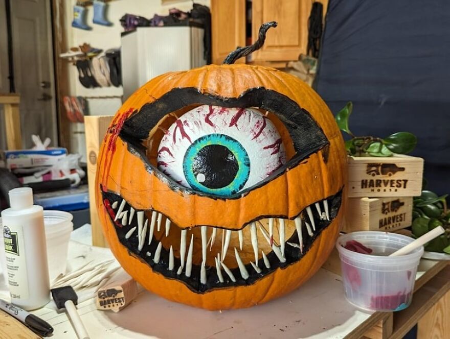 Pumpkin carving featuring a spooky eye and jagged teeth with paint supplies in a home workshop setting.