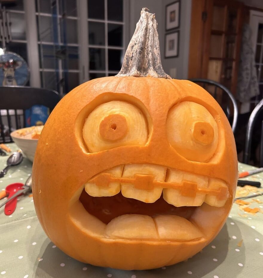Carved quirky pumpkin with exaggerated eyes and braces-style teeth sitting on a table with carving tools nearby.