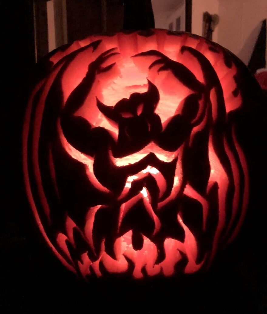 Pumpkin carving of a muscular devil figure surrounded by flames, glowing with an eerie red light at night.