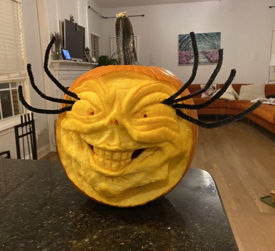 Intricately carved quirky pumpkin with a smiling face and black pipe cleaner spider legs on a kitchen counter.