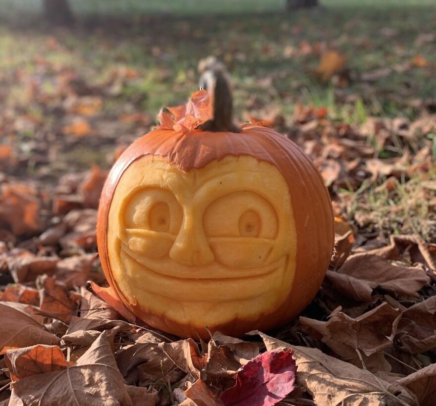 Pumpkin with quirky carved face sitting on the ground surrounded by fallen autumn leaves, showcasing unique pumpkin carvings.