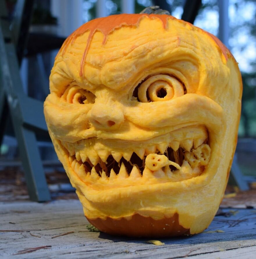 Intricately carved spooky pumpkin carving with sharp teeth and a small figure inside its mouth on a wooden surface outdoors.