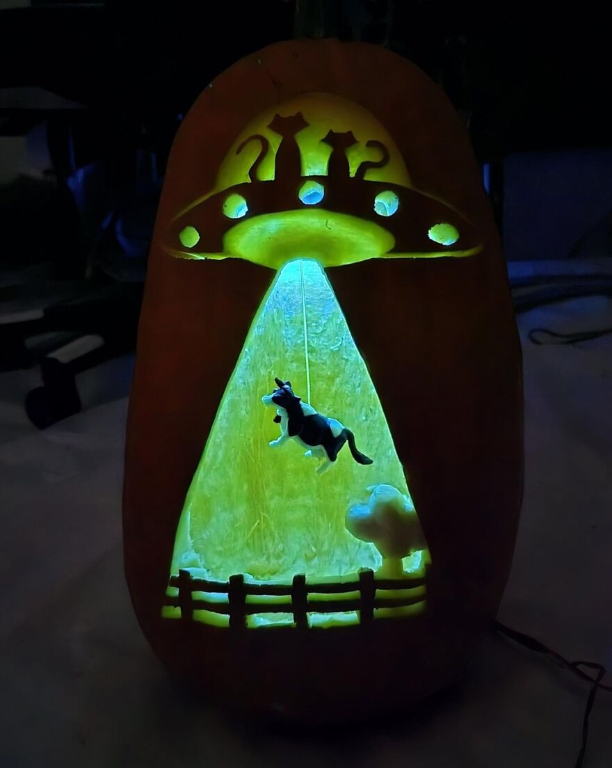 Pumpkin carving of two cats in a UFO abducting a cow with glowing green light and a fence below at night.