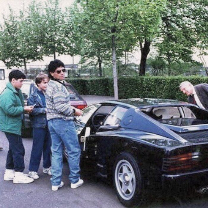 People admiring a black sports car in the '90s, with a mix of casual and curious expressions.