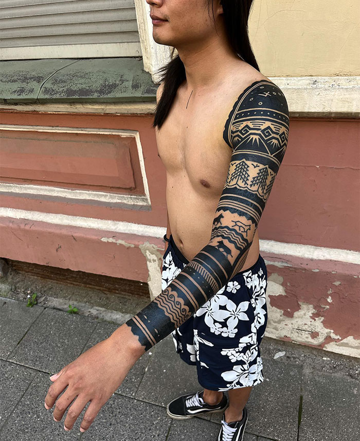 A man with a tribal-inspired full-sleeve tattoo design featuring bold, black geometric patterns and intricate details. Tattoo ideas for men looking for traditional or modern interpretations of tribal art.