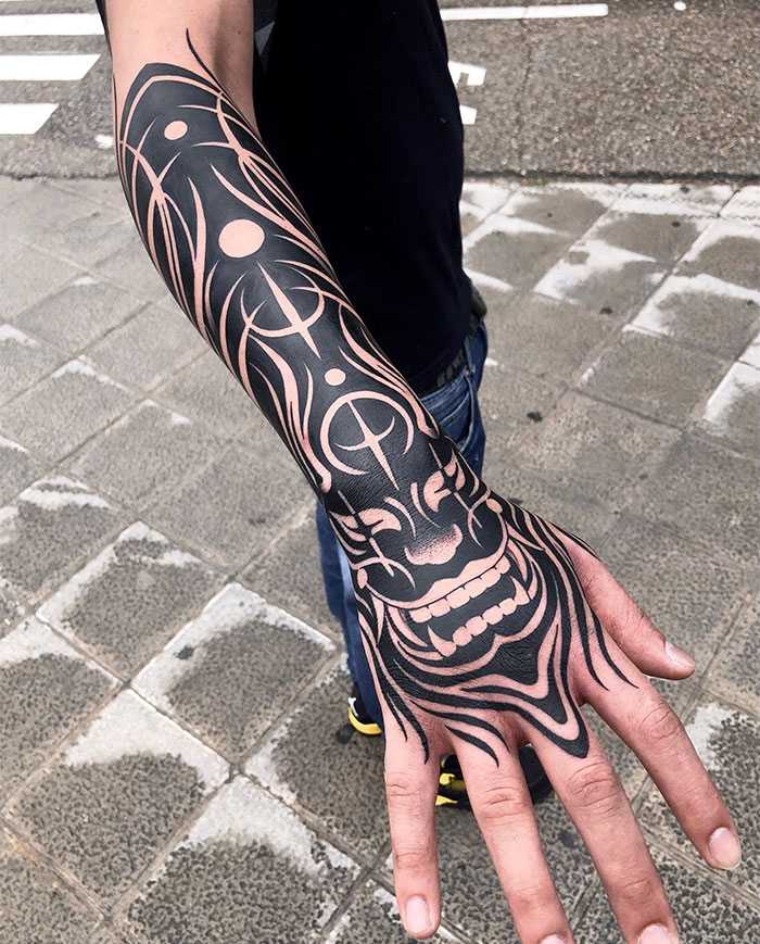 A man showcasing a full-arm tattoo with intricate negative space and bold black ink, featuring a stylized mask and circular motifs. Tattoo ideas for men seeking a combination of traditional and modern tribal designs.
