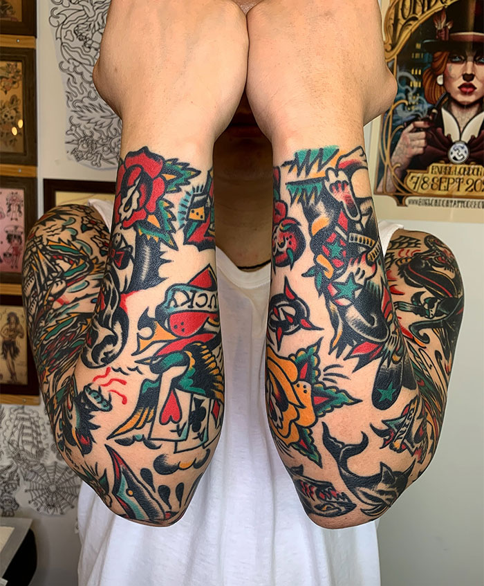  A man displaying both arms fully covered with colorful traditional-style tattoos featuring roses, playing cards, swallows, and bold outlines. Tattoo ideas for men inspired by classic Americana designs with vibrant colors and symbolic motifs.